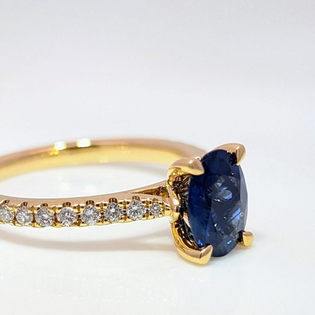 Image depicts one ring facing a south-east angle on a white surface. It's an oval Sapphire wed fit, with diamond set shoulders set in 18k yellow gold and 4 talon claws.